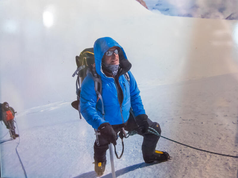 Man in bright blue parka and climbing gear ascends a snowy mountain while he is tied to people ahead of and behind him.