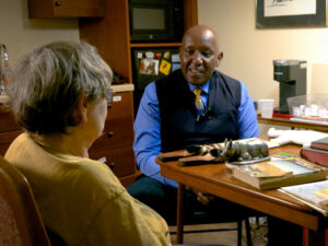 A businessman and senior woman share a table of books and memorabilia from East Africa.