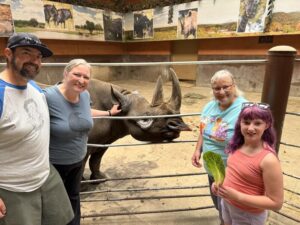 Three adults and a child stand next to a rhinoceros behind a fence. One is petting the rhino behind her ear.