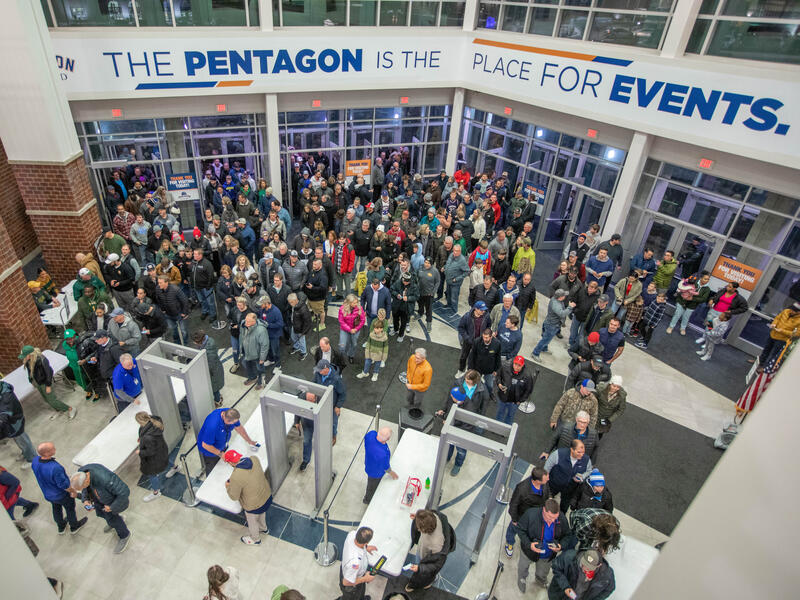 Overhead view of fans streaming into a sports venue lobby with metal detectors and security personnel. A banner reads "The Pentagon is the place for events."