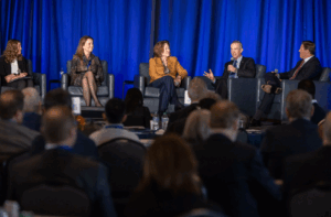 Three businesswomen and two businessmen sit on stage in front of a blue curtain. One of the men speaks into a microphone.
