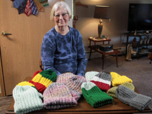 Senior woman sits behind a table full of knitted stocking caps.
