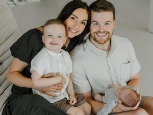 A mom and dad sit together while holding their toddler and newborn baby.