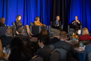 Businesspeople sit on stage, listening to one man talking into a microphone.