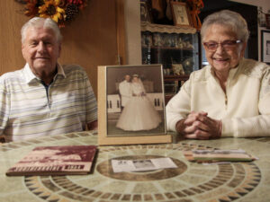 Senior couple poses at their kitchen table with their framed wedding photo.