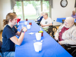 Young woman speaks with senior person across a dining room table at a nursing home. Other seniors chat in the background.