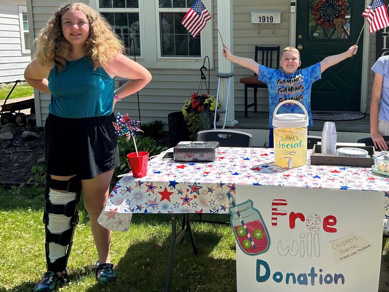 A girl in a knee brace and a boy waving American flags sell lemonade on a sunny day. The sign on their table says "free will donation."