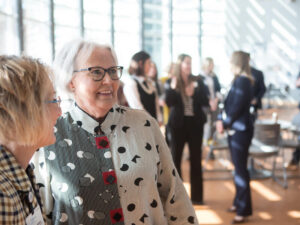 Happy, mature businesswoman visits with others in a sunny lobby. Groups of people stand talking behind them.