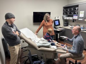 Boy with one skinny leg and one average leg sits in an exam room with doctor at his feet and parents at his sides.