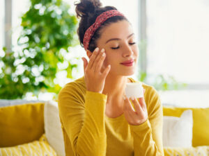 Young woman with eyes closed applying face cream.