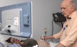 Older doctor scans belly of pregnant patient while they both look at a computer screen.