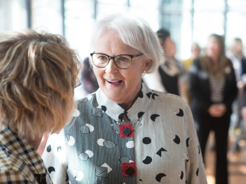 Happy, mature businesswoman visits with others at an event in a sunny lobby.