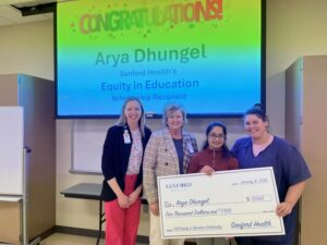 Four people hold a giant check in front of a screen that says "Congratulations! Arya Dhungel, Sanford Health Equity in Education scholarship recipient"