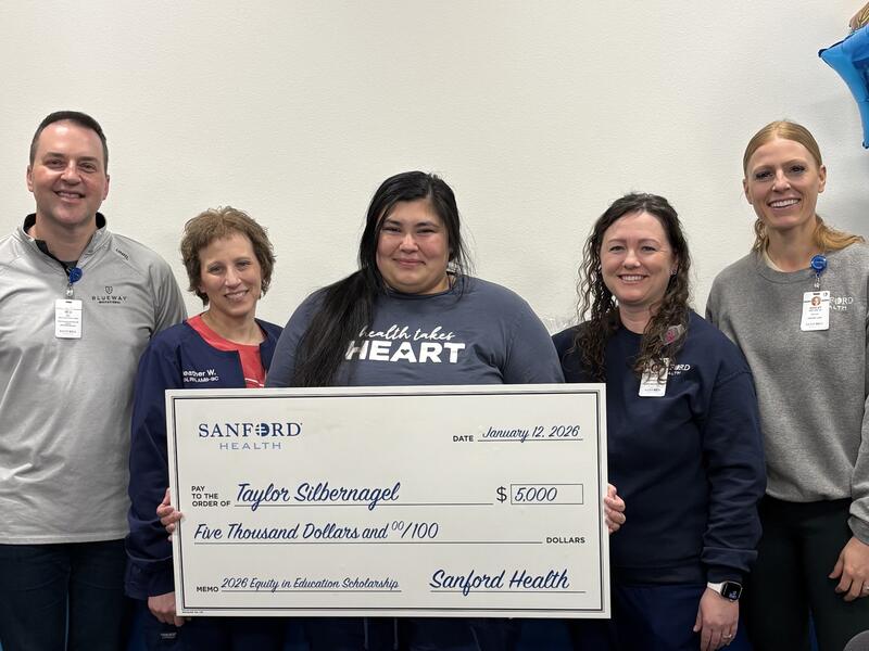 Five people hold a giant check for Taylor Silbernagel's Sanford Health Equity in Education Scholarship.