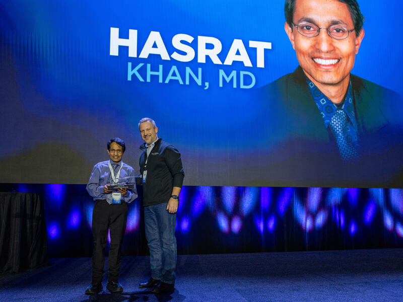 Two men onstage in front of a photo of Hasrat Khan, MD.