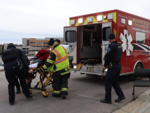Emergency responders wheel a man on a guerney into the back of an ambulance while a police officer stands guard with a safety cap on his rifle.