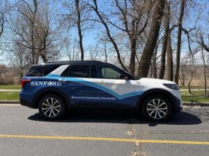 SUV marked Sanford Health "Bismarck Community Paramedic" drives on a tree-lined road.