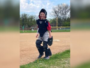 10-year-old boy in catcher's uniform smiles on outdoor baseball field.