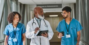 Screenshot from HealthTech. Three doctors, two in scrubs and one in a white coat and suit, talk as they walk down the hall.