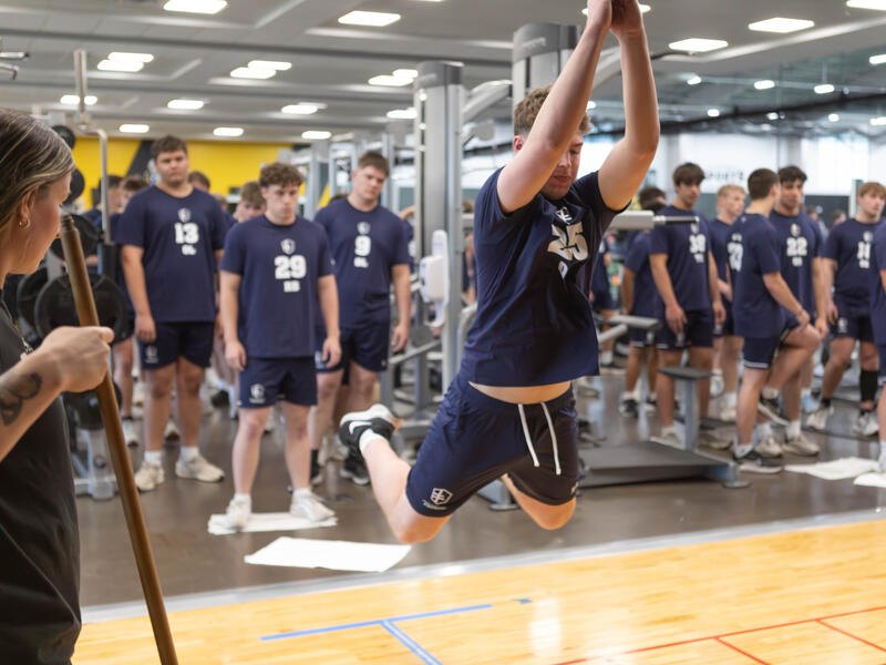Student athletes line up to take turns as one boy jumps across the floor.