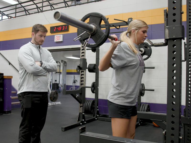 Trainer watches student athlete lift weights in gym.