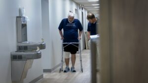 Down a hallway, a man with a knee bandage uses a walker with a physical therapist helping him.