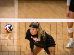 College volleyball player focuses on net ahead of her with teammates and another volleyball in background.