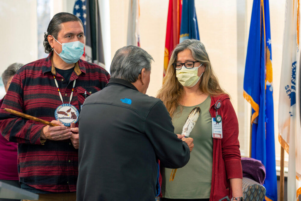 Two Sanford Health workers in surgical masks stand at the front of a flag-draped room, one holding a ceremonial pipe, while a Native American advisor waves an eagle feather.